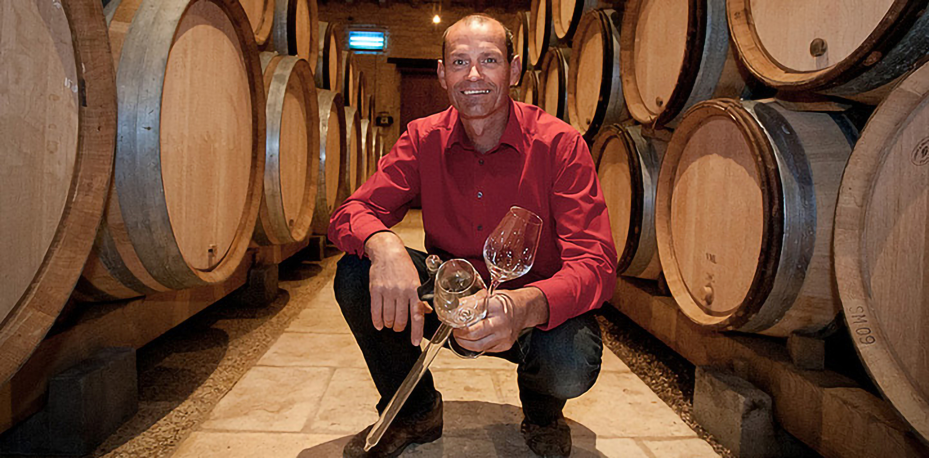 Jean-Philippe Fichet kniet mit einer Pipette und zwei Gläsern im Keller vor Weinfässern und lächelt in die Kamera