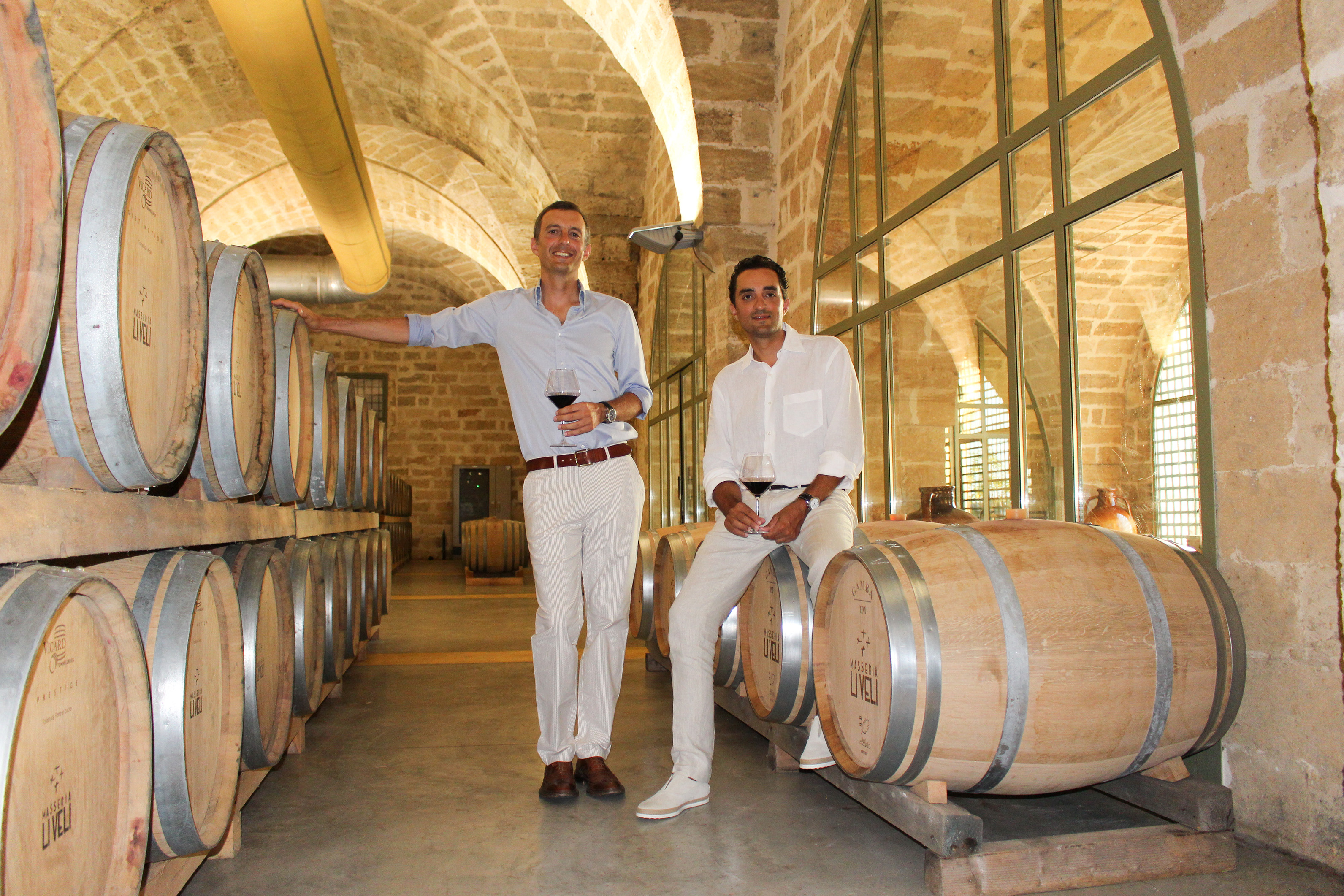 die beiden Winzer von Masseria Li Veli sitzen auf Weinfässeren in deren Weinkeller mit Gewölbe und trinken Rotwein