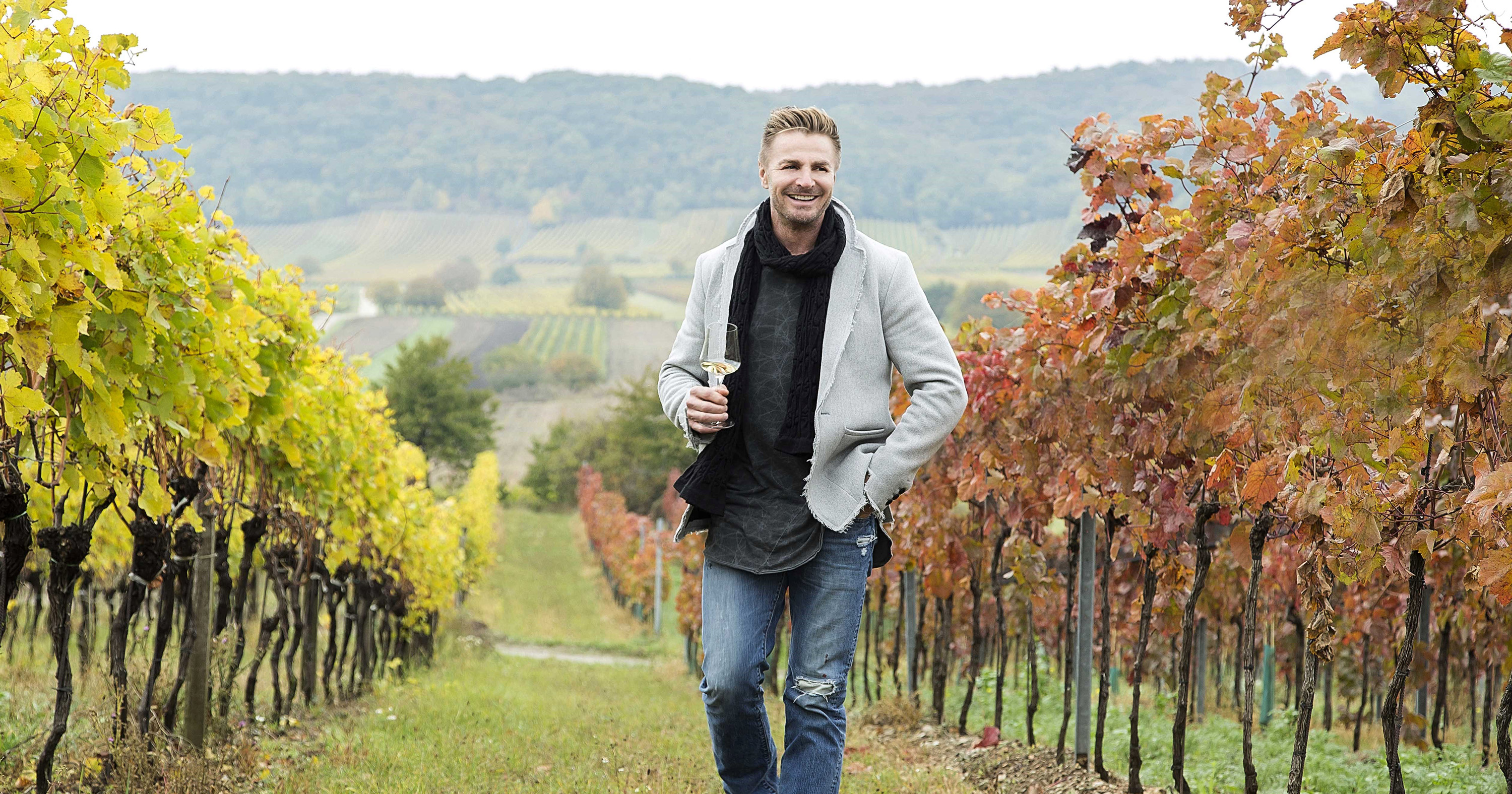 Leo Hillinger spaziert an einem Herbsttag durch den Weingarten mit einem Glas Weißwein in der Hand